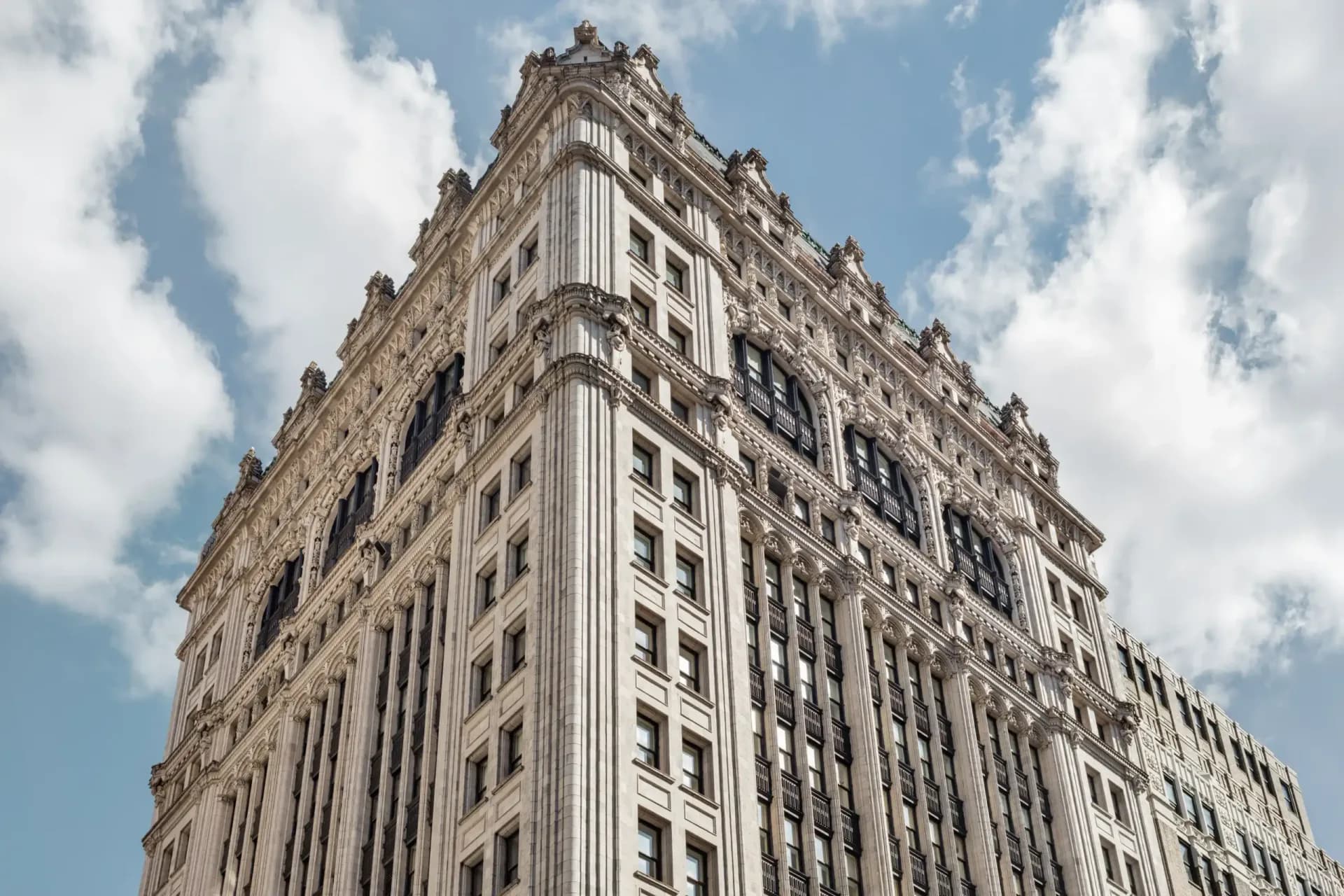 Low-angle view of an ornate stone skyscraper facade with carved details against a blue sky with clouds.