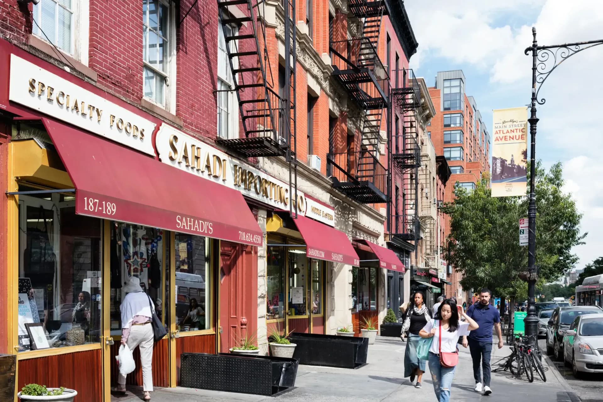 Busy city sidewalk lined with red awnings and storefronts including Sahadi's specialty foods shop, pedestrians walking past, in a Historic Districts Council gallery.