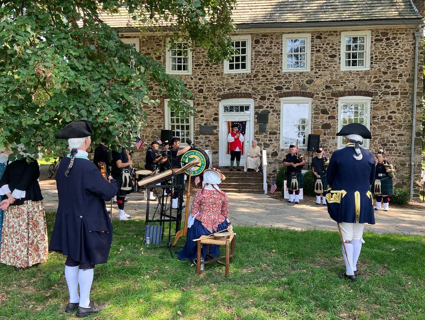 Stone historic house exterior with ivy, surrounded by costumed interpreters in 18th-century military and civilian dress conducting outdoor demonstrations on the lawn.