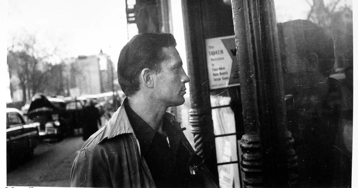 Black and white photograph showing man in profile wearing dark jacket with striped collar, standing near pole with urban street and parked cars visible behind him.