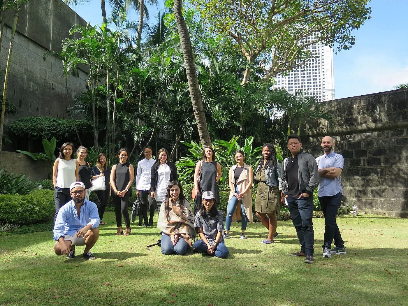 Group of approximately 20 people standing and sitting on a lawn in front of stone walls and tropical vegetation, with modern buildings visible in the background.