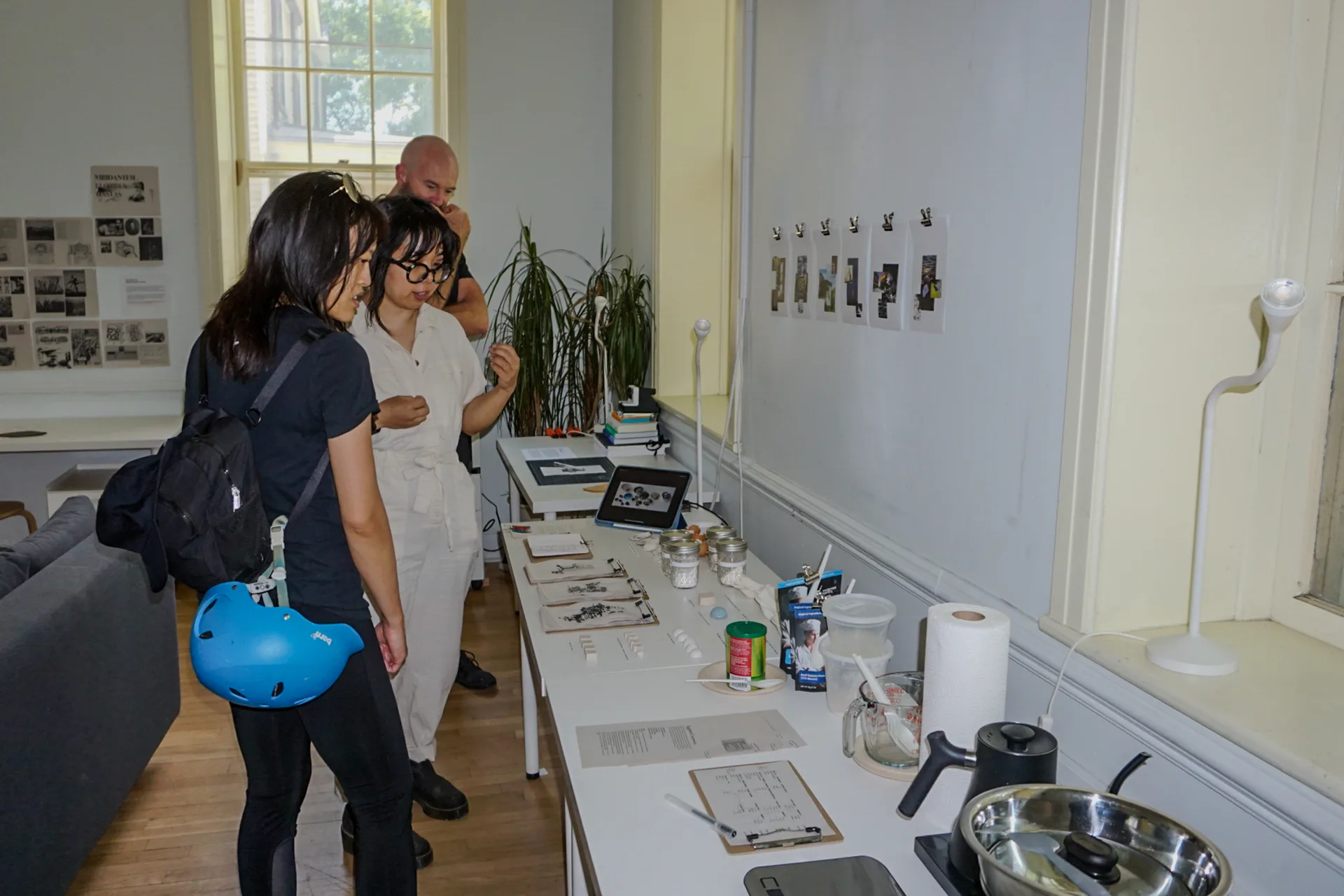 Gallery interior with white walls displaying small framed works, visitors examining a table-mounted exhibition of objects and materials.