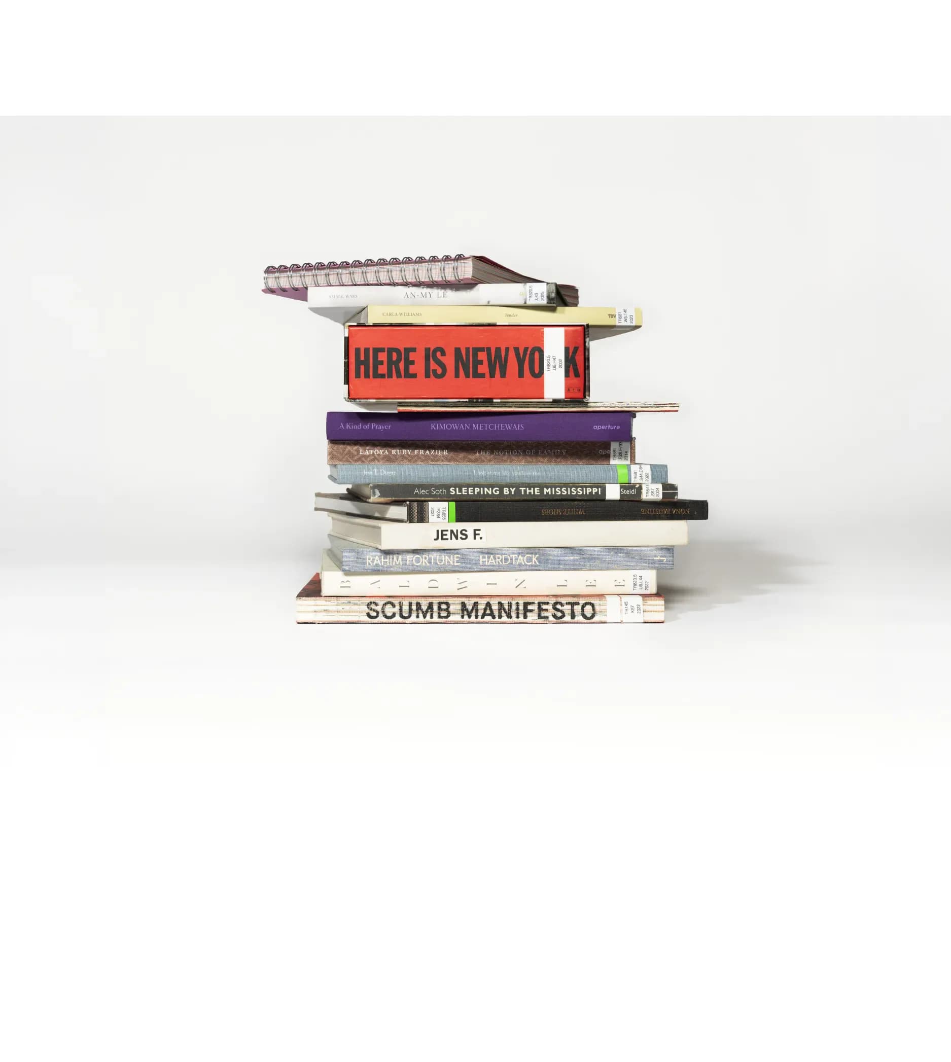 Stacked books photographed against white background, including titles 'Here Is New York,' 'Scumb Manifesto,' and others, arranged as a sculptural composition.