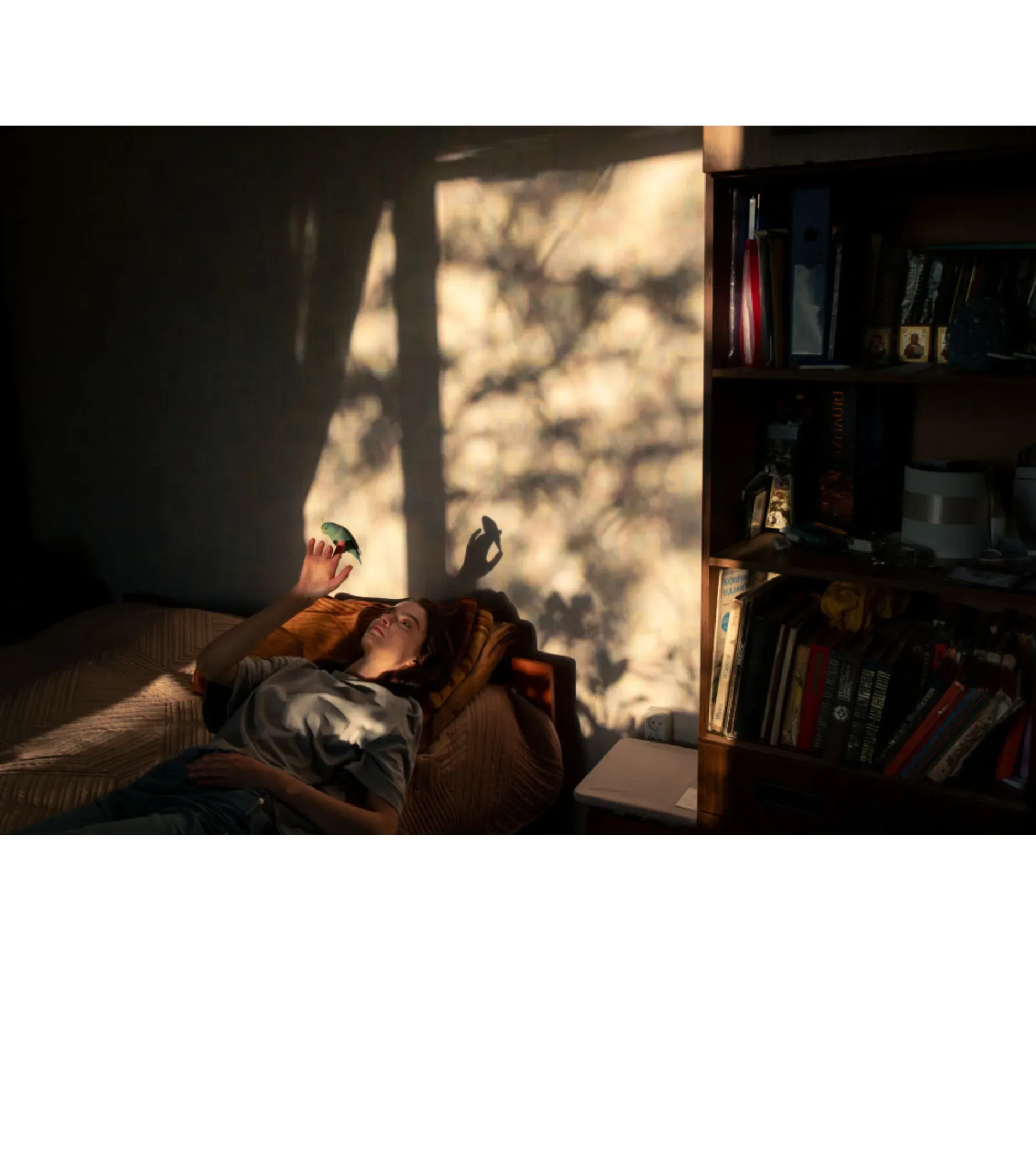 Black and white photograph of a person sitting on a couch in dramatic sunlight streaming through a window, with bookshelves visible on the right side.