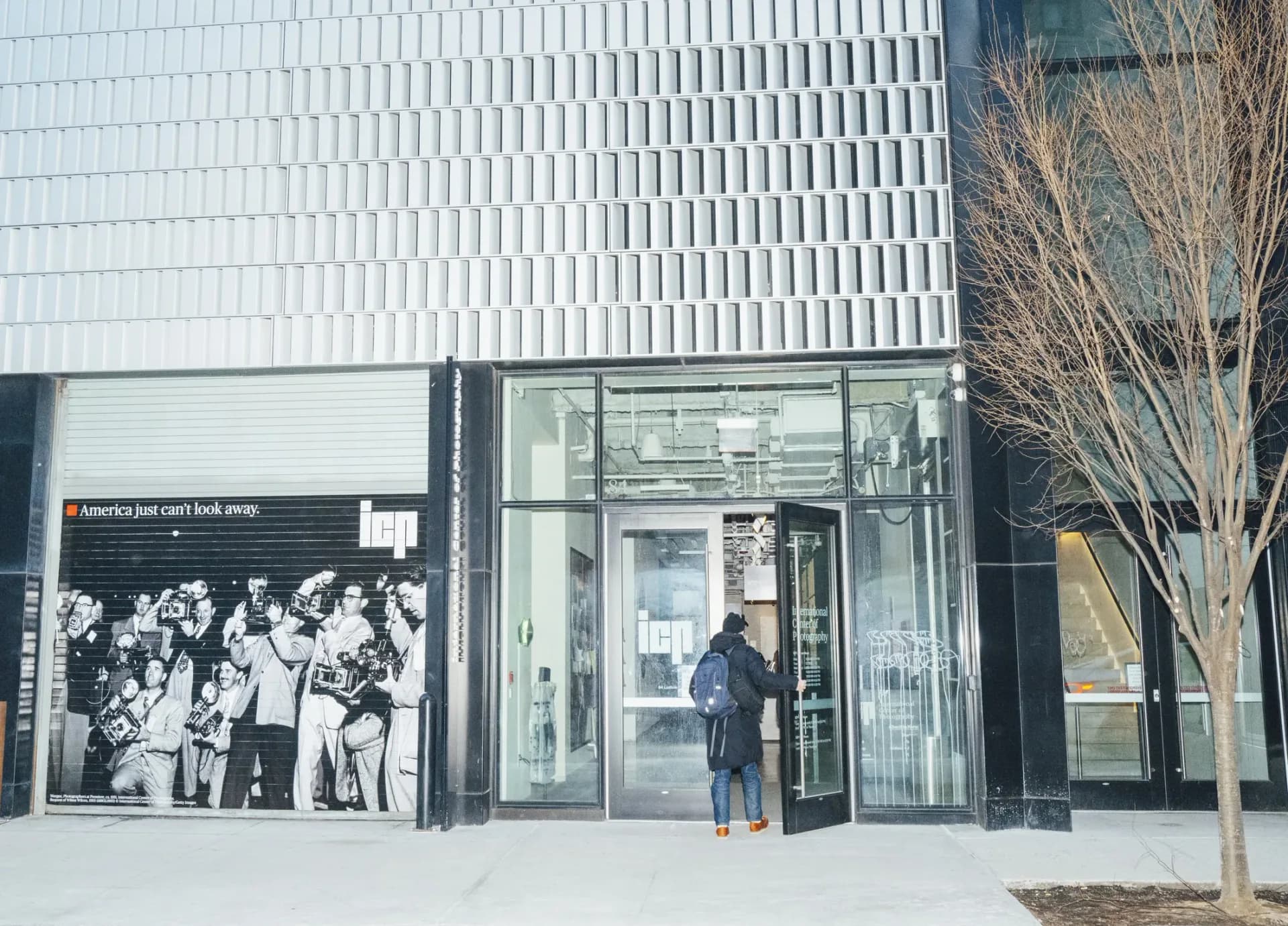 Exterior storefront photograph of a modern gallery building with white ribbed facade, glass entrance doors, and a person entering; bare tree visible on right.