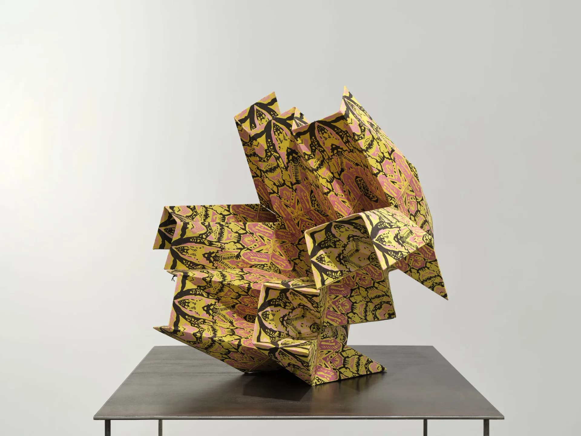 Abstract folded sculpture with yellow and green patterned fabric featuring botanical motifs and brown accents, mounted on a gray platform.