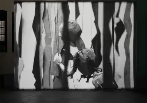 Black-and-white video still or projection showing abstracted human figures among draped vertical fabric or translucent forms, creating a fragmented, ethereal composition.