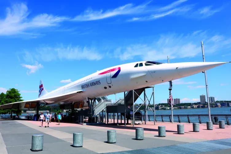 Concorde supersonic aircraft displayed outdoors as a museum exhibit with visitors on a plaza, waterfront visible in background.