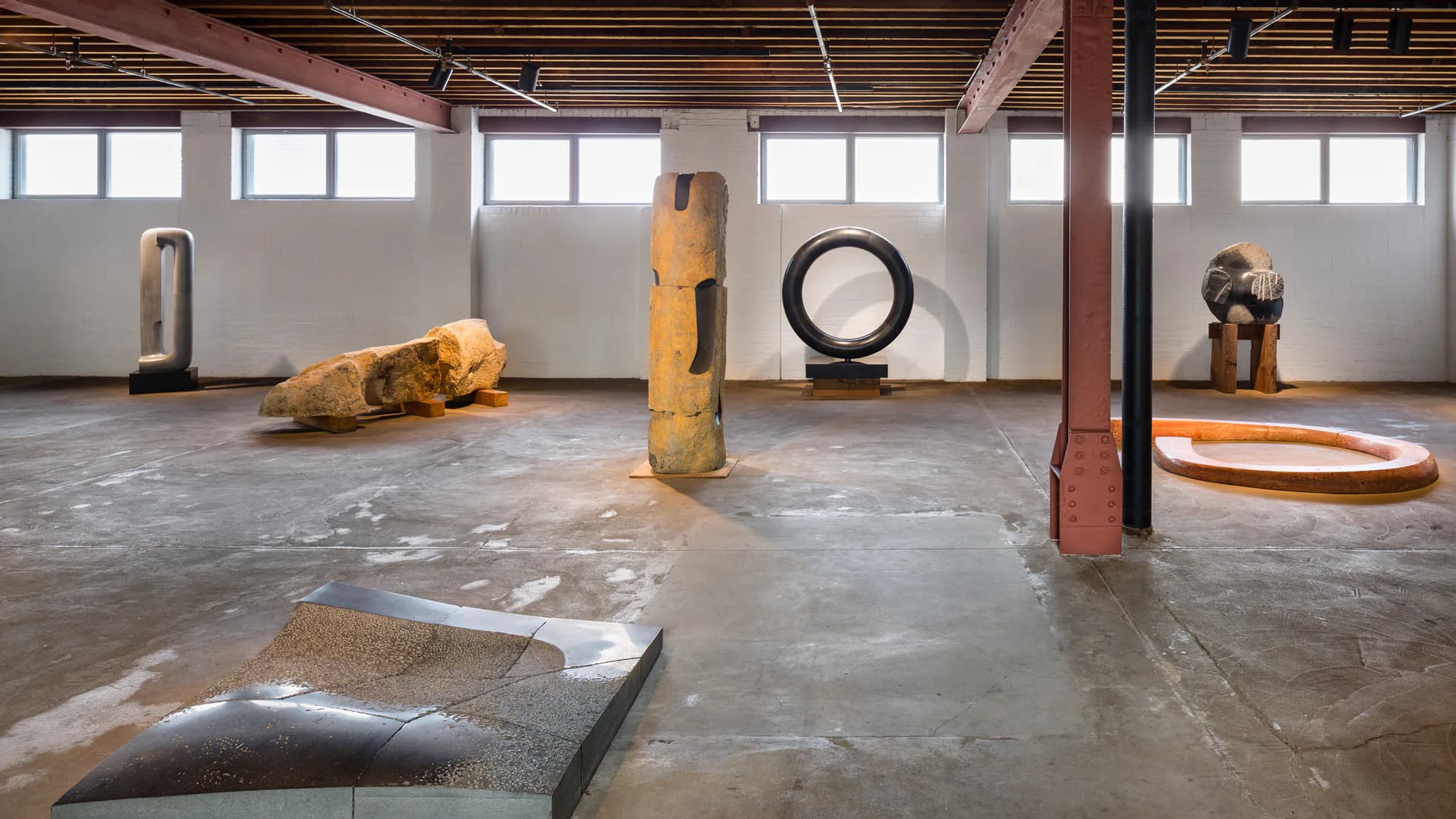 Industrial gallery space with concrete floor, large windows, exposed red structural columns, displaying various abstract wooden and metal sculptures and forms throughout.