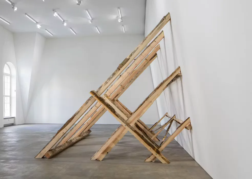 White gallery interior with wooden beam sculpture leaning against wall, track lighting overhead, polished concrete floor.