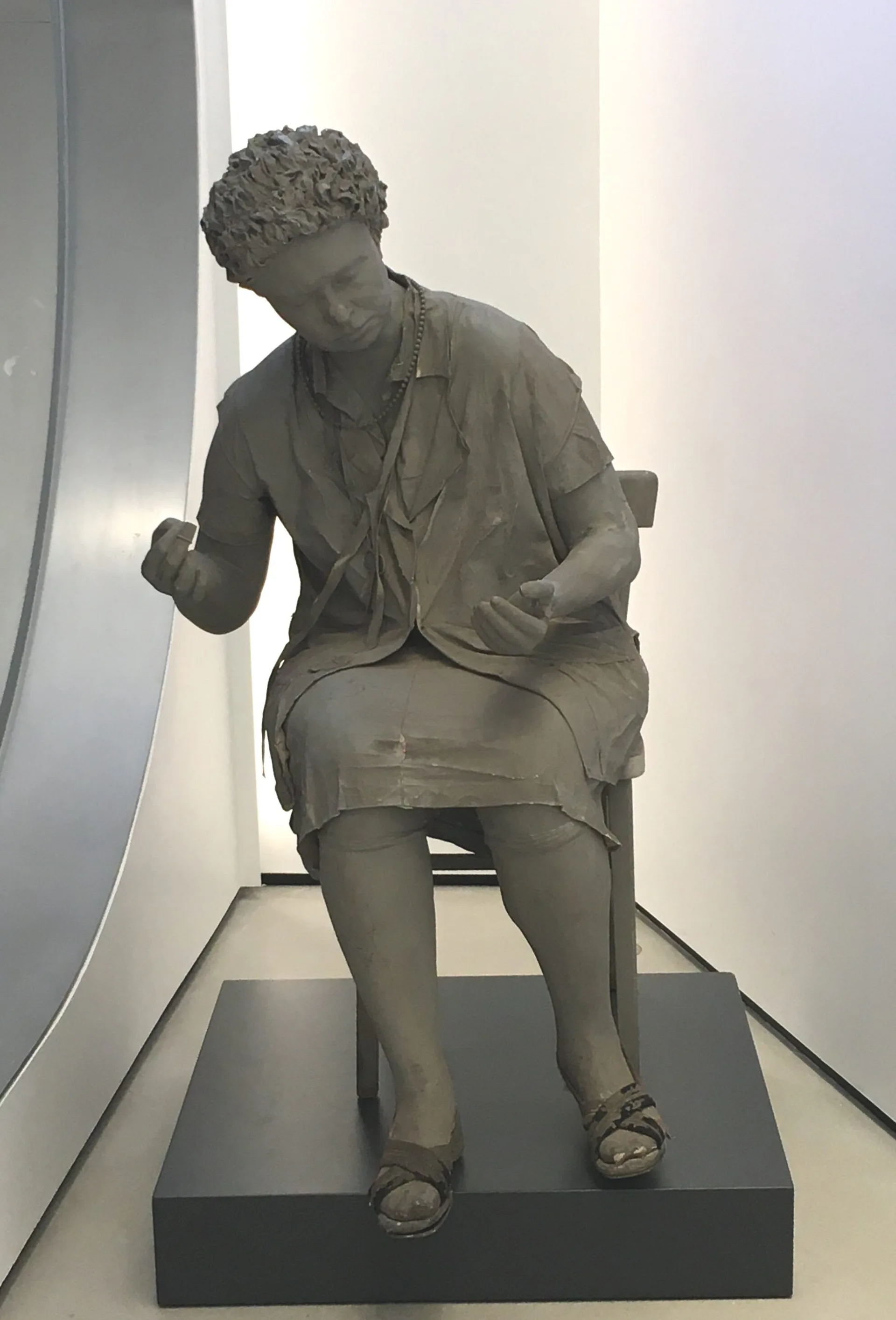 Stone sculptural figure of a seated woman in contemplative pose, displayed on a black pedestal in a minimalist gallery setting.