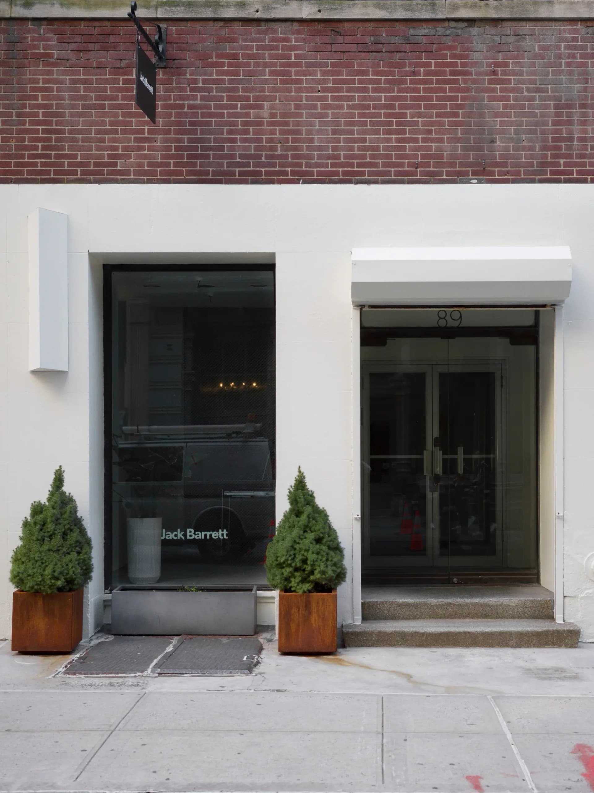 Storefront of Jack Barrett gallery with white facade, glass doors marked 89, and two potted evergreen trees flanking the entrance