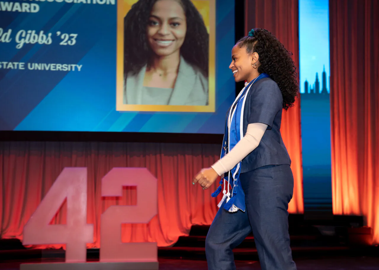 Woman in dark blazer speaking on stage with portrait and 'Gibbs '23' text visible on screen behind her.