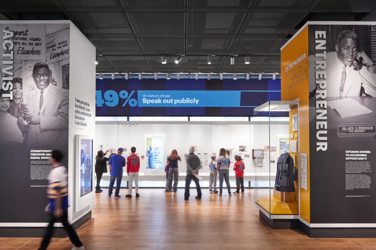 Museum exhibition interior with large banners featuring historical portraits and statistics, visitors browsing displays.