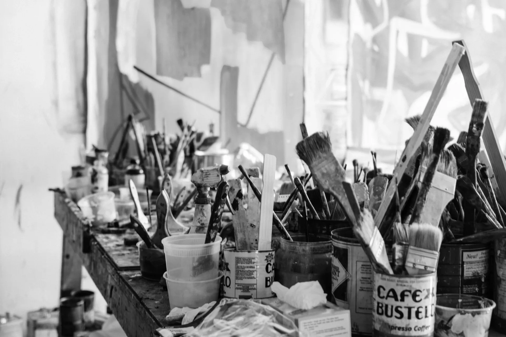 Black and white photograph of artist's studio workspace with containers of brushes, paint tubes, and art supplies on table with draped fabric.