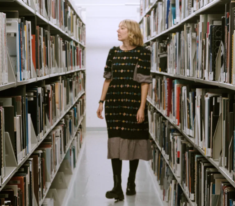 Library or archive interior showing tall shelving units with books and media on both sides of a center aisle, with a woman in a patterned dress walking through the space.