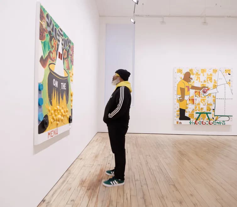White gallery space with light wood flooring, person viewing two colorful artworks mounted on white walls with artwork featuring yellow tones and figurative imagery.