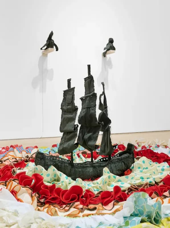 Installation artwork featuring dark fabric castle-like structure surrounded by colorful crumpled textile and suspended figurative elements.