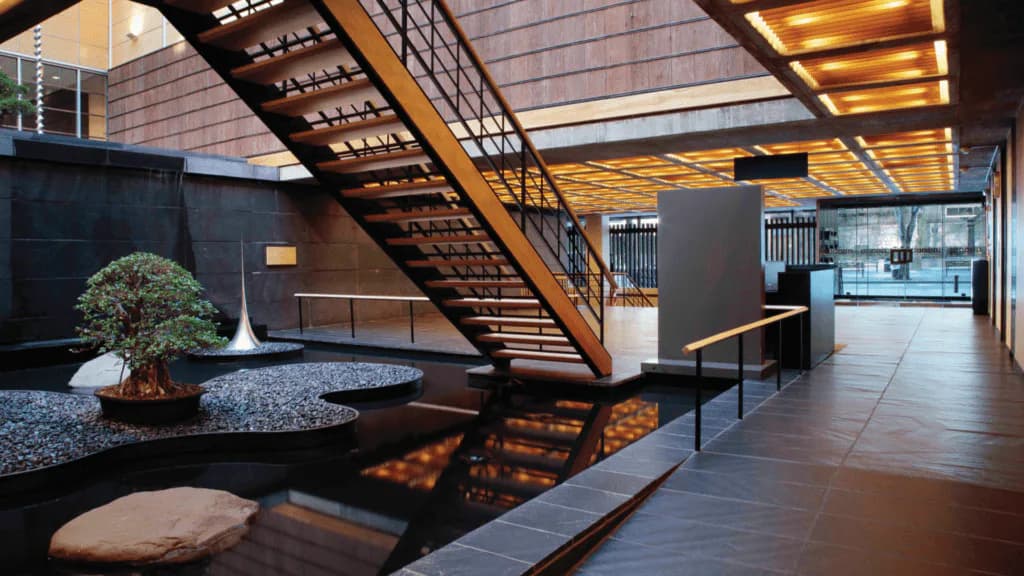 Modern gallery interior with wooden staircase, polished floors, bonsai tree on dark platform, and warm overhead lighting at Japan Society.