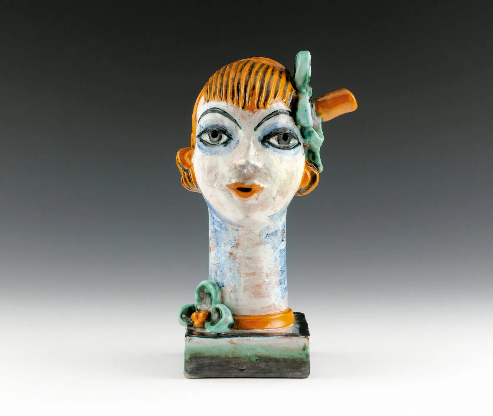 Ceramic sculptural head with orange-striped hair, white face, blue eyes, and orange and green accents, mounted on rectangular base.