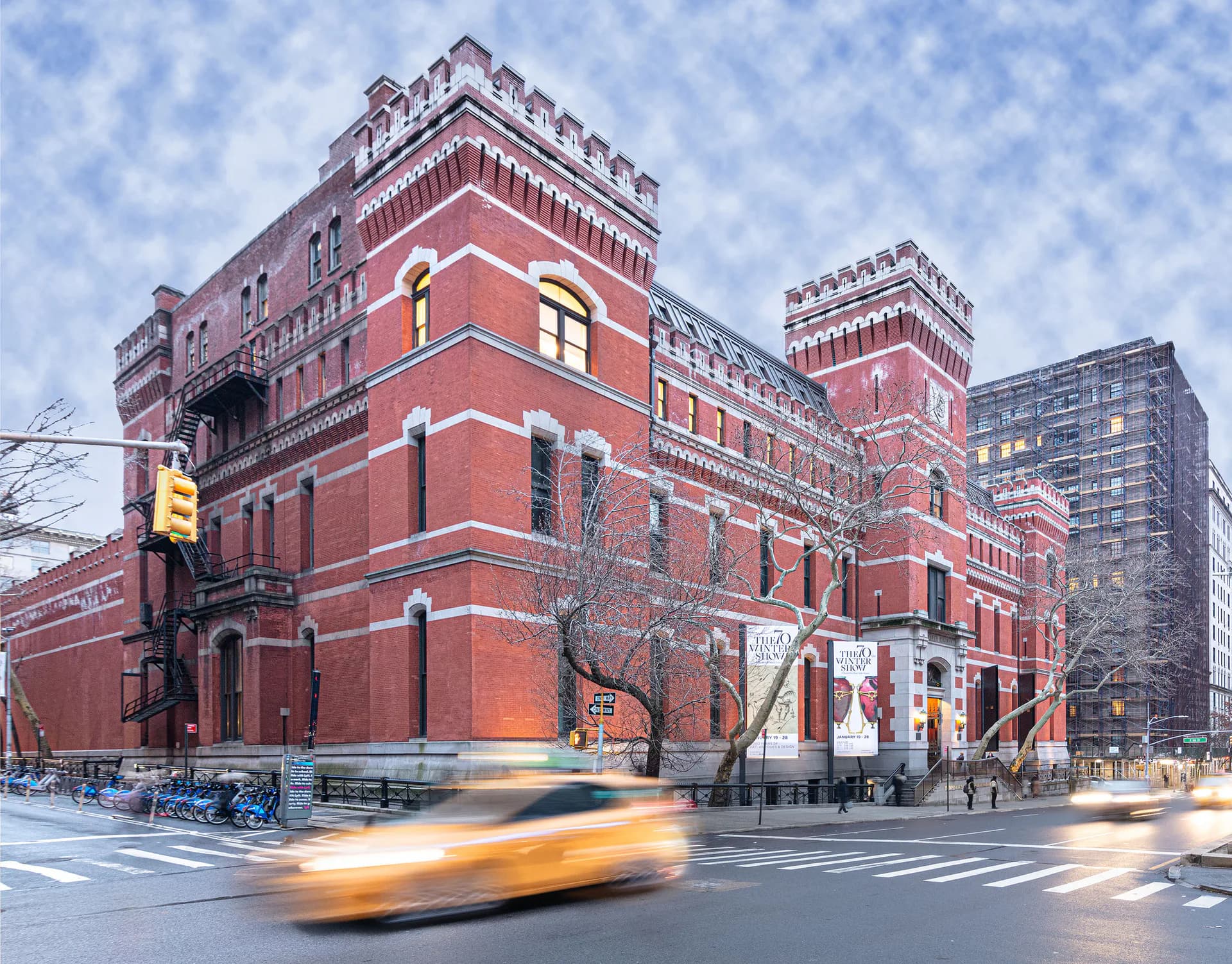 Exterior photograph of Park Avenue Armory, a red brick building at night with illuminated yellow taxi in foreground and snow on ground.