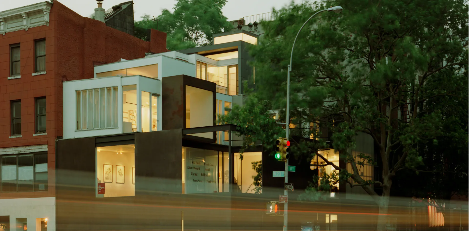 Modern glass and dark-paneled building exterior at dusk with warm interior lighting, adjacent to red brick structure and mature trees.