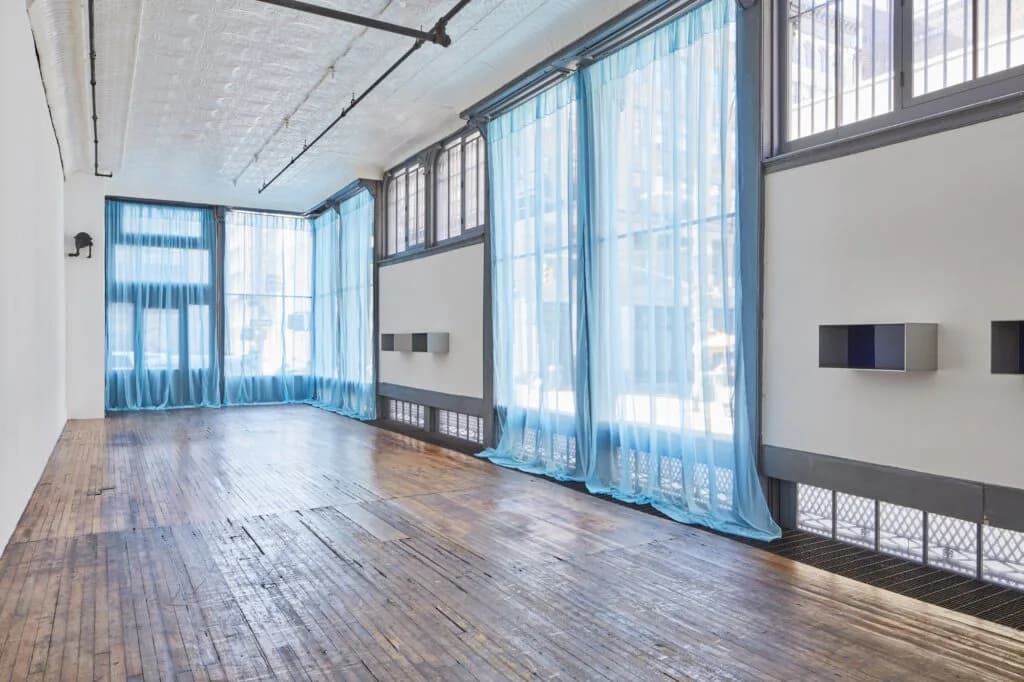 Judd Foundation gallery interior with blue sheer curtains, wooden flooring, industrial windows, and minimal wall-mounted elements.