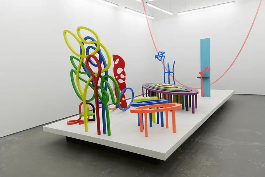 White gallery platform displaying colorful sculptural installation with interlocking looped forms in neon colors, nested furniture structures, and curved architectural elements.