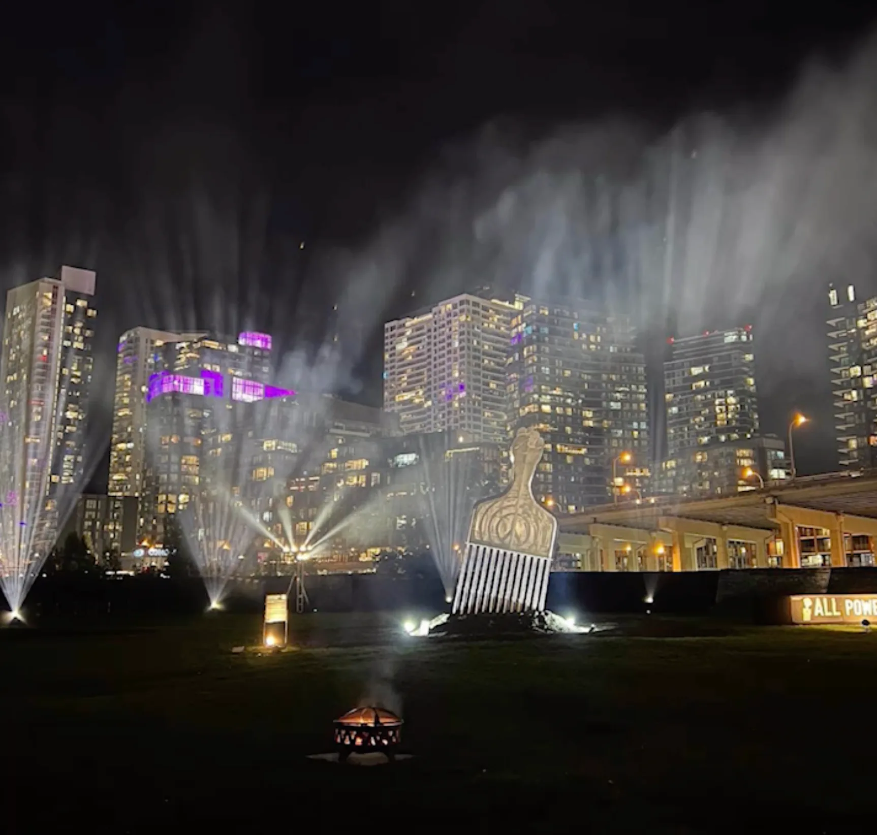 Night photograph of monumental outdoor light installation with architectural sculptures illuminated against downtown cityscape and high-rise buildings.
