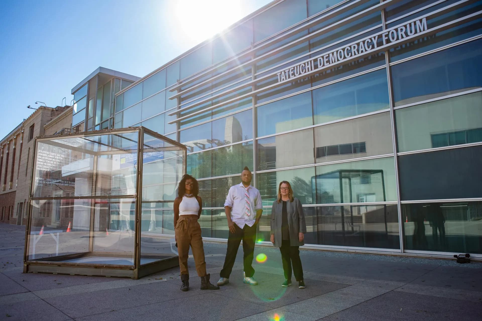 Outdoor plaza in front of Chicago History Museum Forum building with three people standing near a transparent sculptural installation in sunlight.