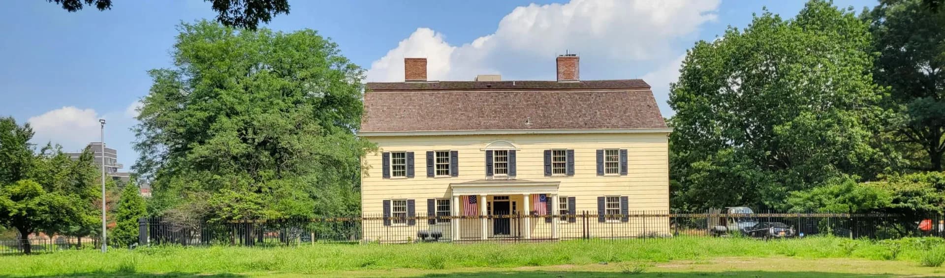 Exterior of King Manor, a two-story yellow colonial house with black shutters and twin chimneys, surrounded by green lawn and trees.