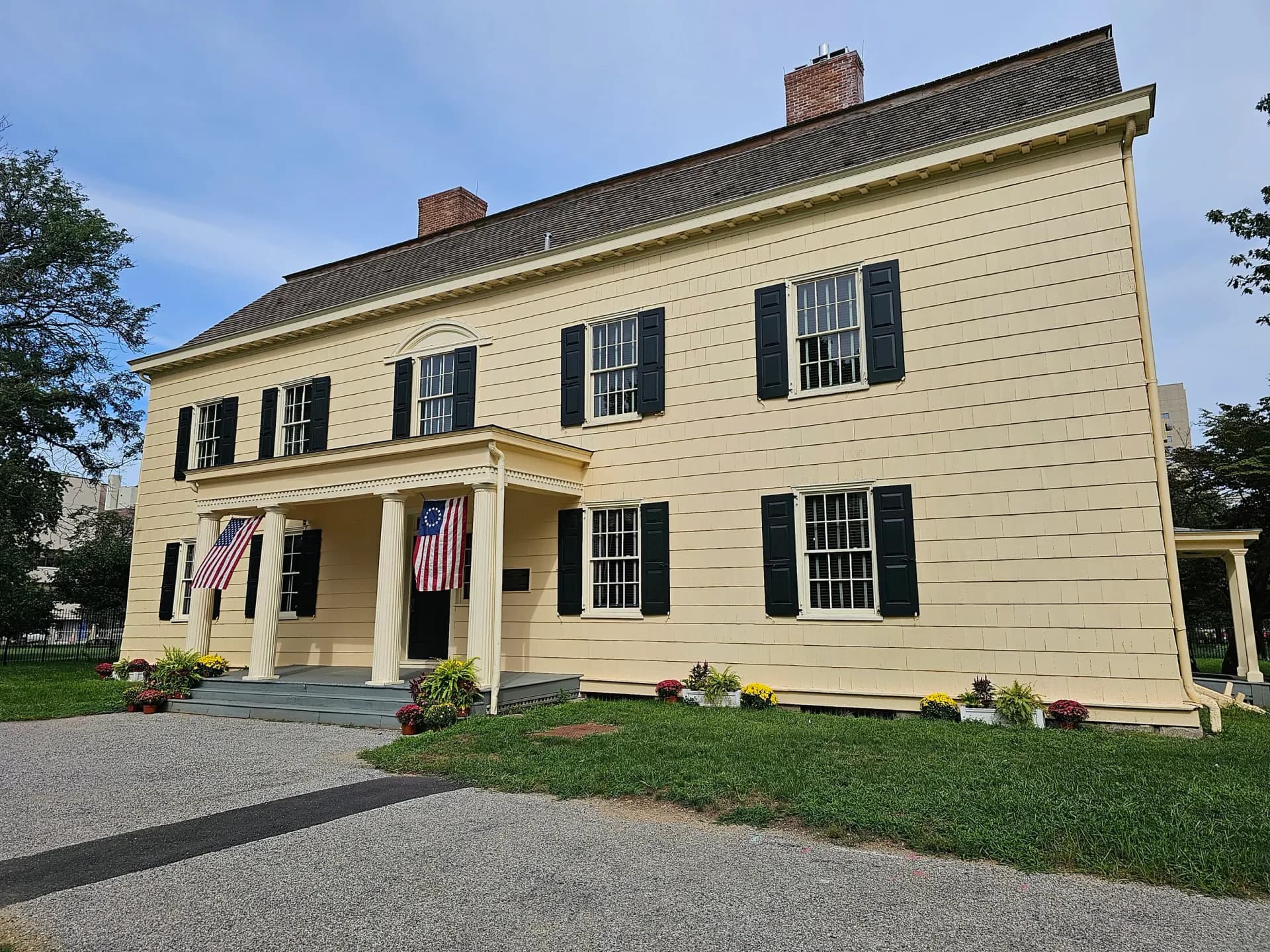 Two-story cream-colored colonial house with black shutters, front porch, American flag, and manicured landscaping.