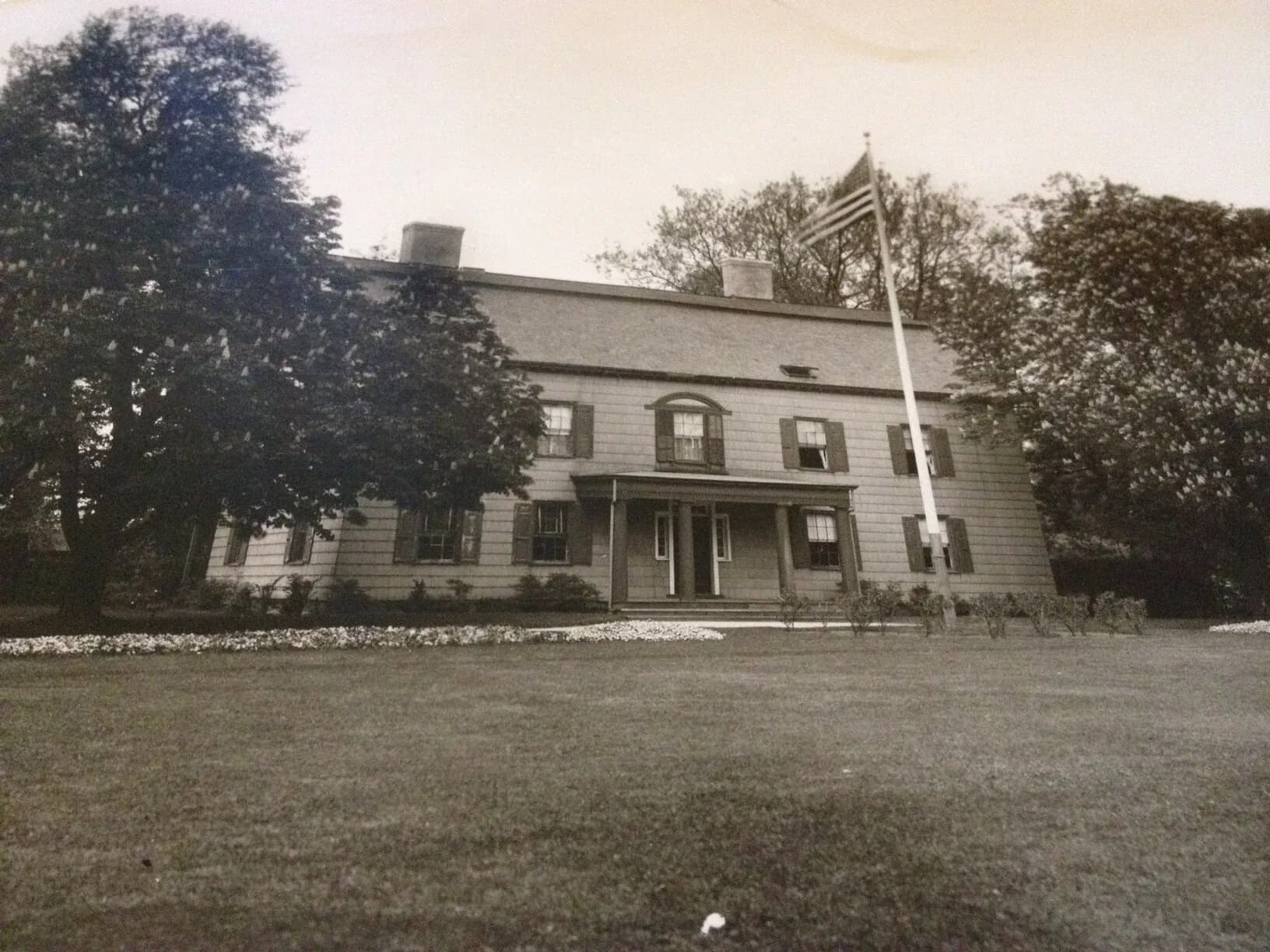 Vintage sepia-toned photograph of a two-story colonial house with large front porch and mature shade trees.