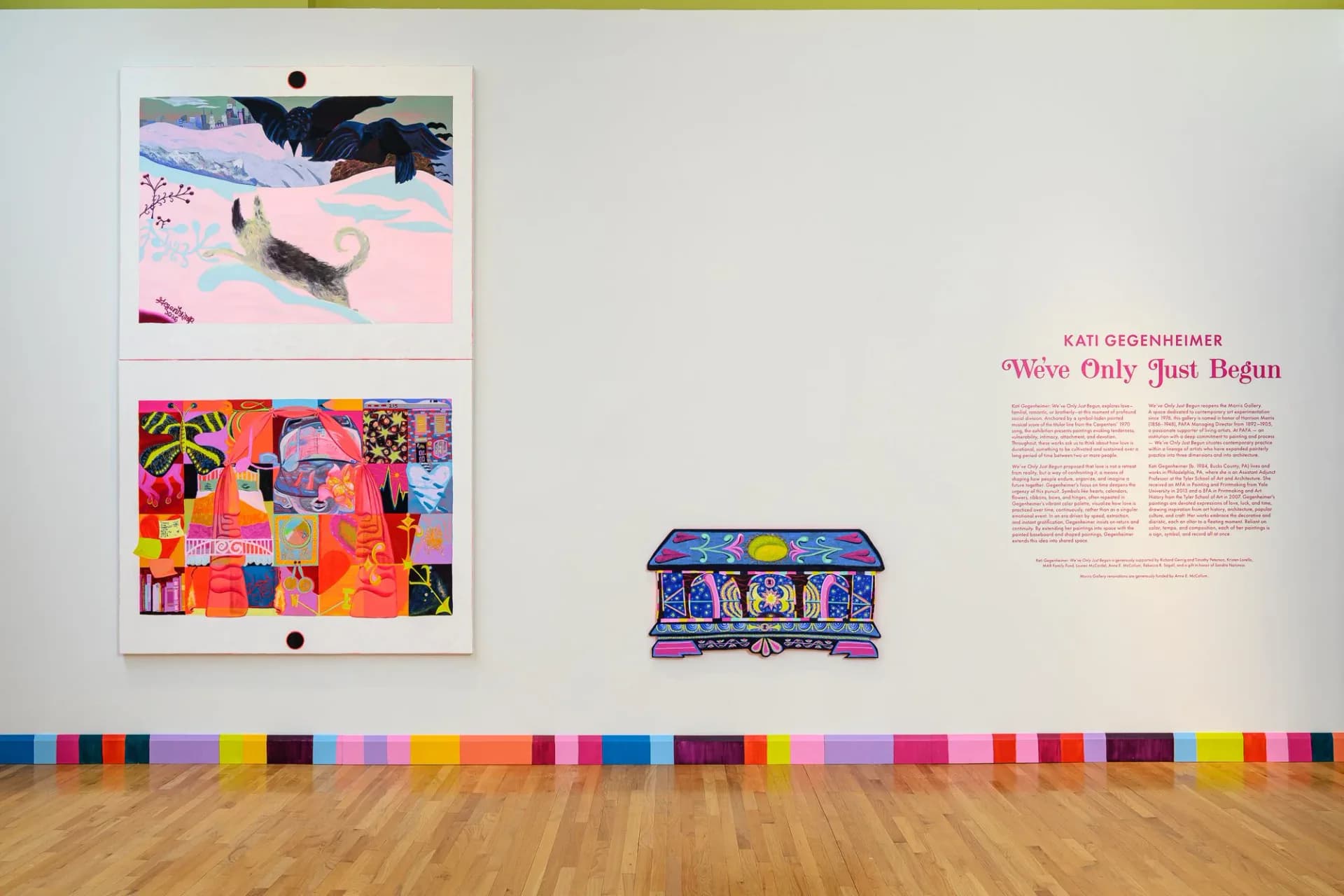 Gallery wall displaying two framed colorful artworks, a small sculptural piece, and descriptive text panel, with a rainbow-colored floor stripe running across the wooden base.