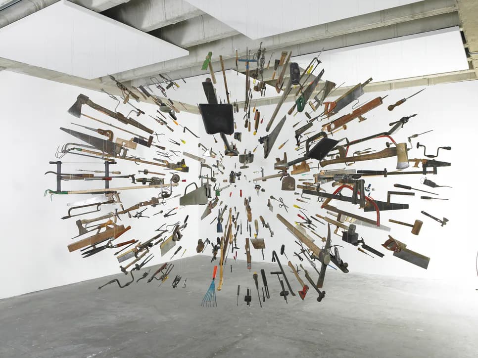 Large-scale sculptural installation of tools radiating outward in three-dimensional burst formation from central point, suspended in gallery space with industrial ceiling.