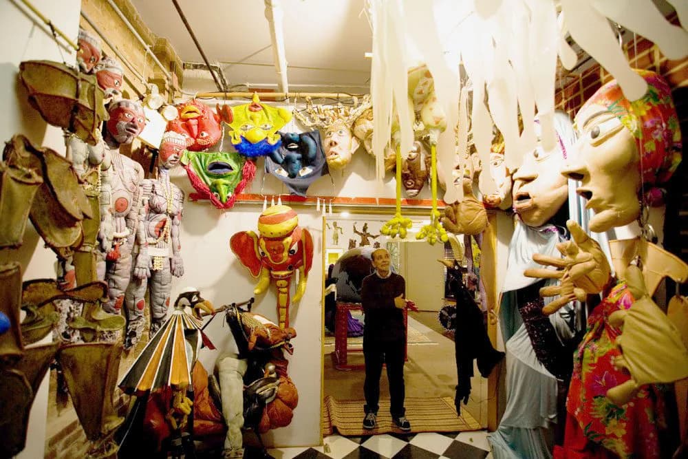 La MaMa gallery space filled with colorful costume pieces, masks, and theatrical props hanging from ceiling and displayed on walls, visitors viewing installations.
