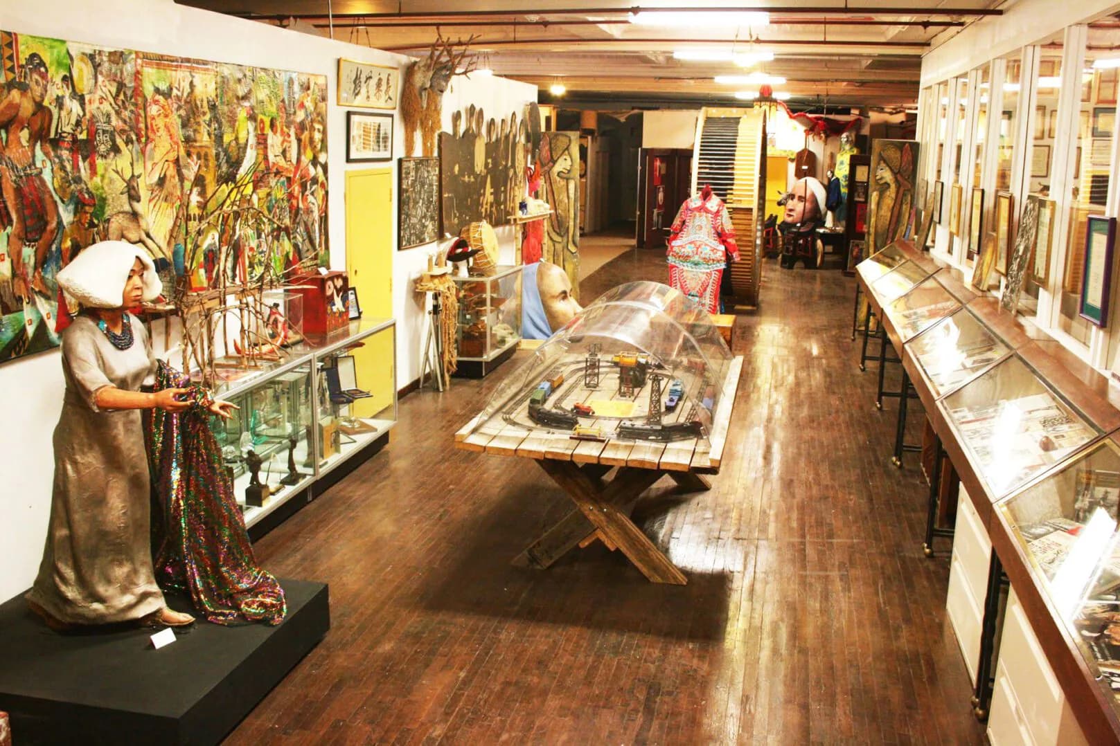 La MaMa gallery interior with wood flooring, display cases, mounted artworks on walls, figurative sculptures, and visitors examining exhibits.