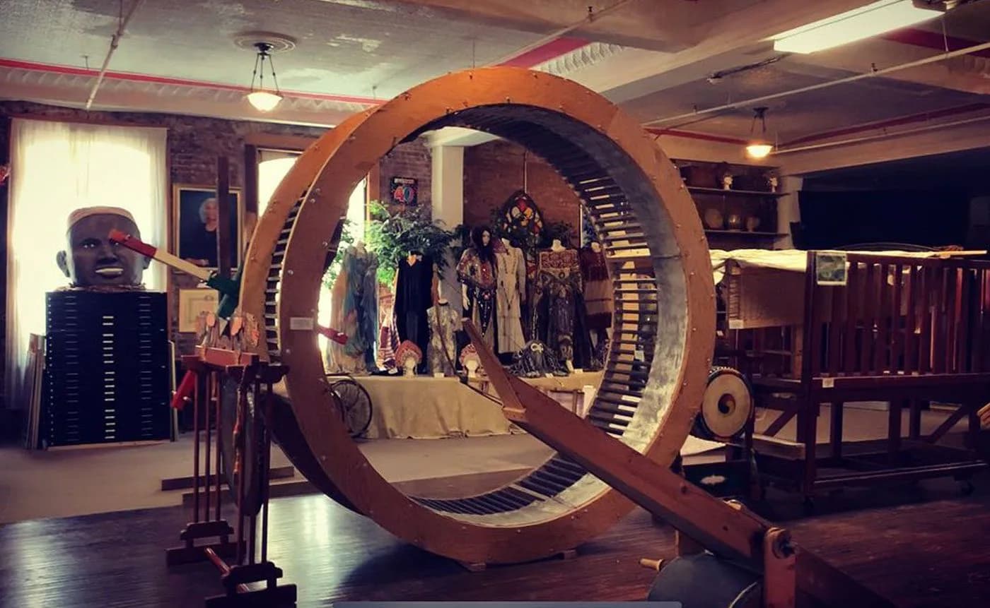 La MaMa gallery space featuring large wooden circular installation with figures inside, vintage furniture, and theatrical costumes displayed throughout.