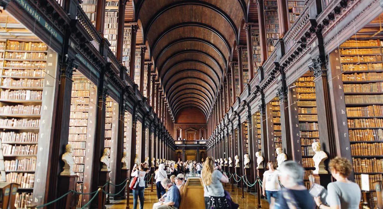 Long corridor of a historic library with soaring barrel-vaulted ceiling, tall wooden bookshelves lining both sides, ornate columns, and visitors walking through the symmetrical passage.