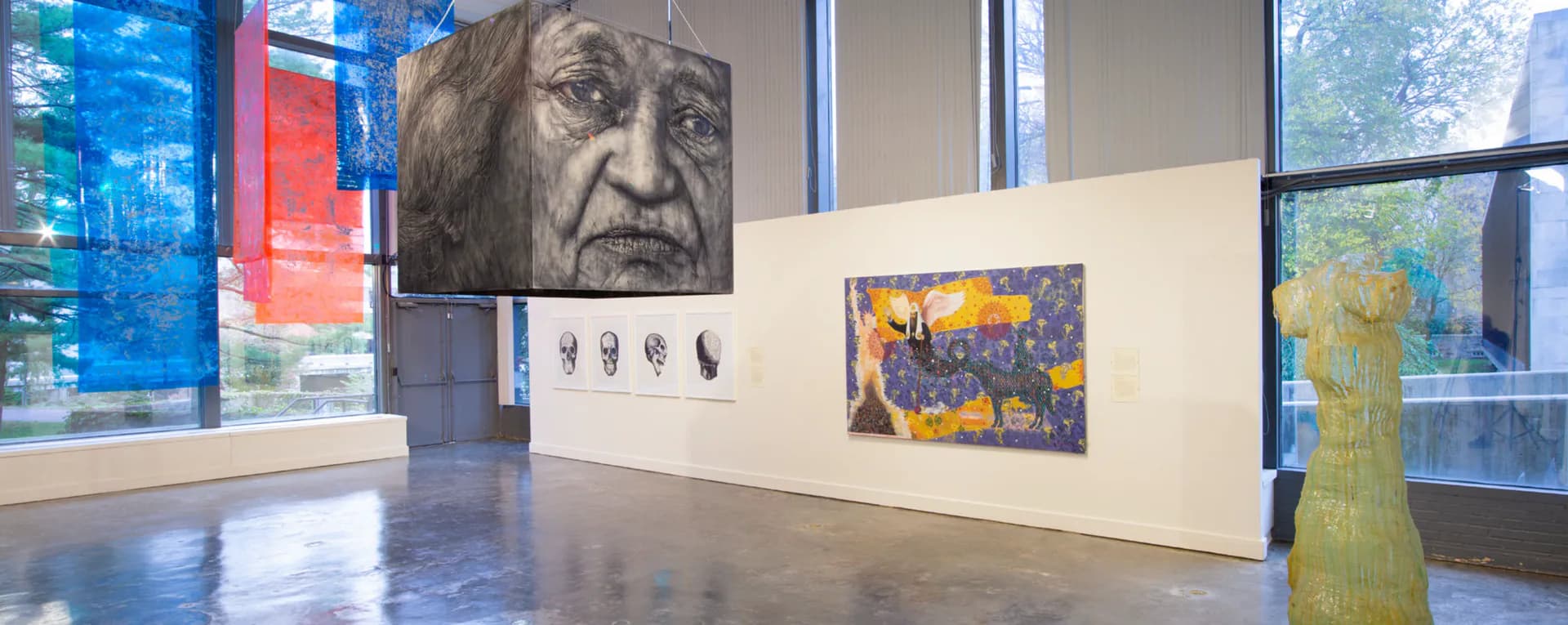 Gallery interior at Lehman College Art Gallery with white walls displaying a large portrait head, smaller artworks, and a sculptural form, with windows overlooking greenery.