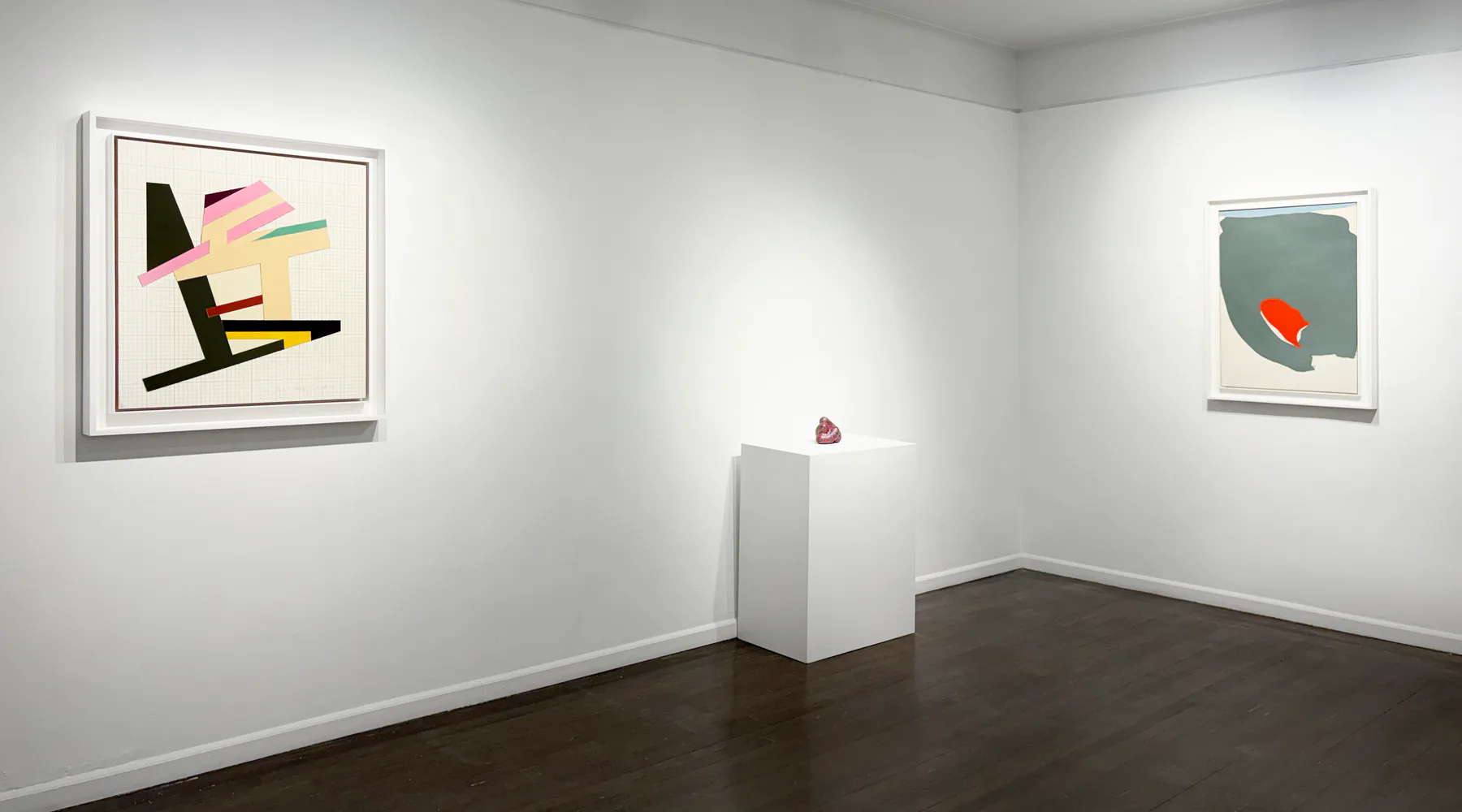 Gallery interior with white walls and dark floor; two framed abstract artworks hang on walls flanking a white pedestal with sculpture.