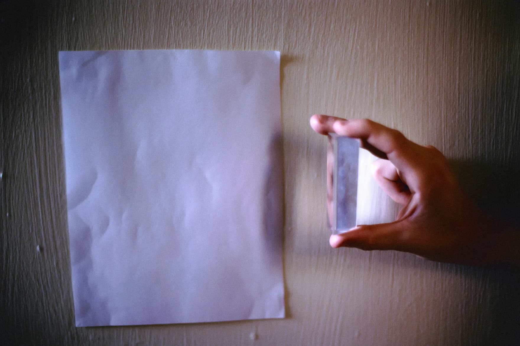 Hand holding a translucent prism or glass object against a white paper background, creating subtle light refraction effects on the surface.