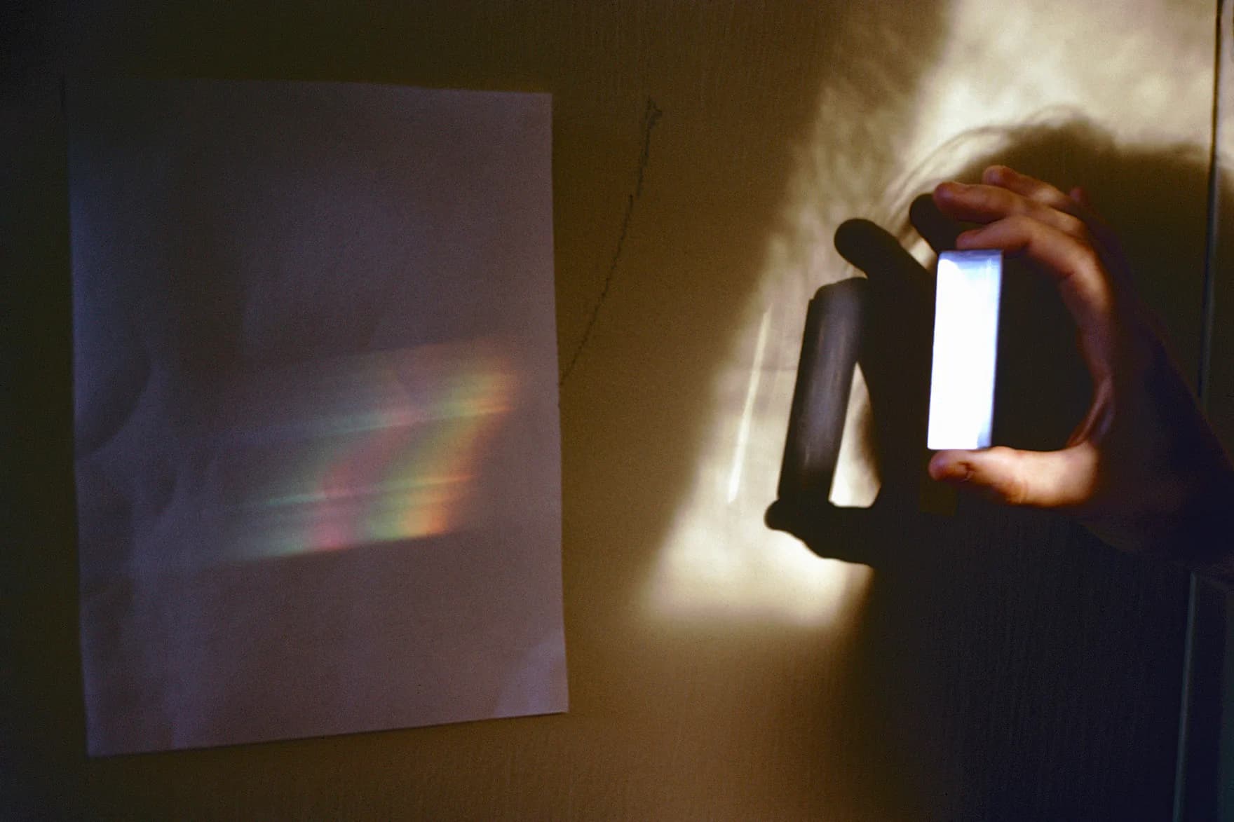 Hand holding a reflective or translucent object projecting a rainbow spectrum onto a white wall in a dark interior space.