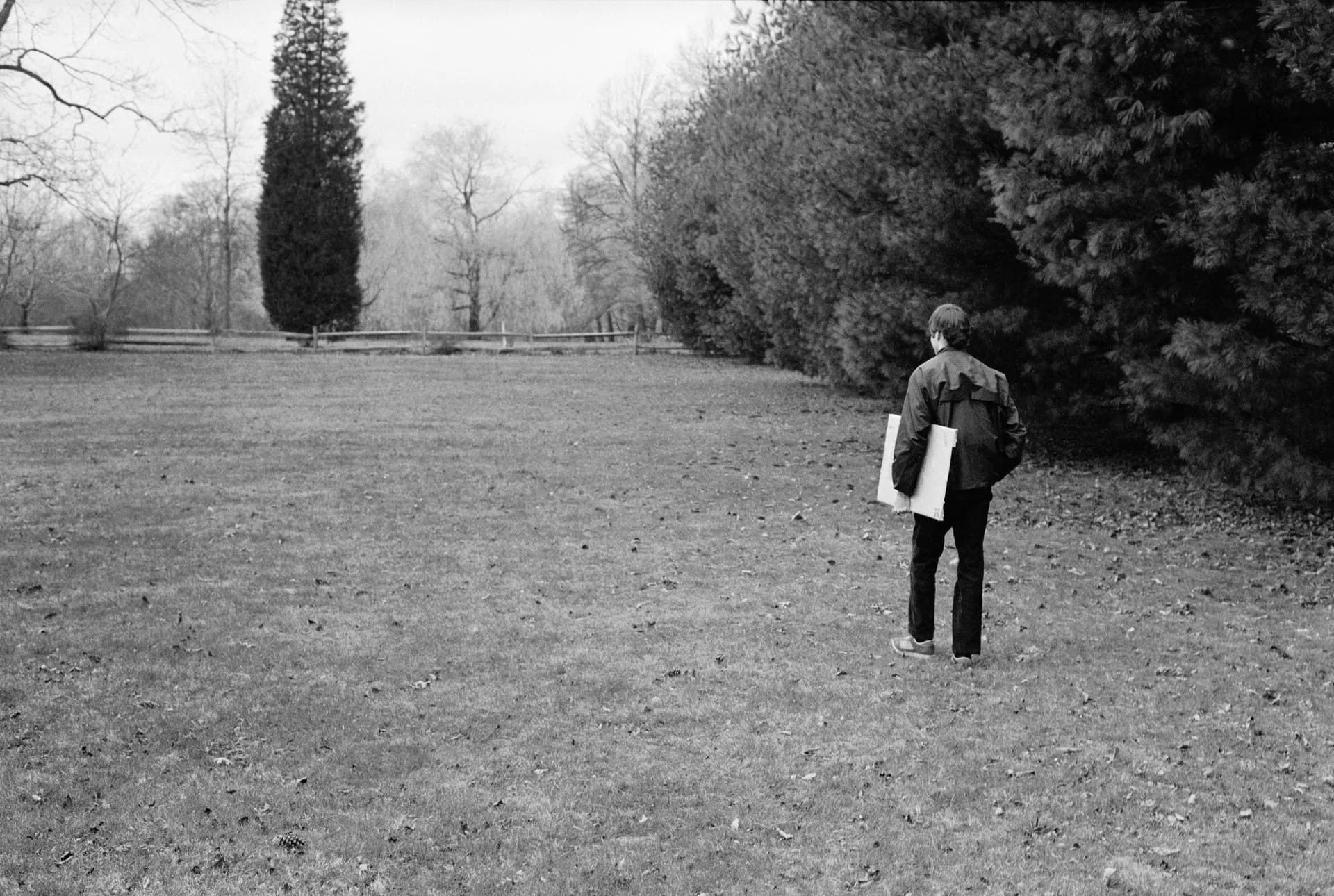 Black and white photograph of a solitary figure viewed from behind holding a white rectangular object in a bare winter landscape.