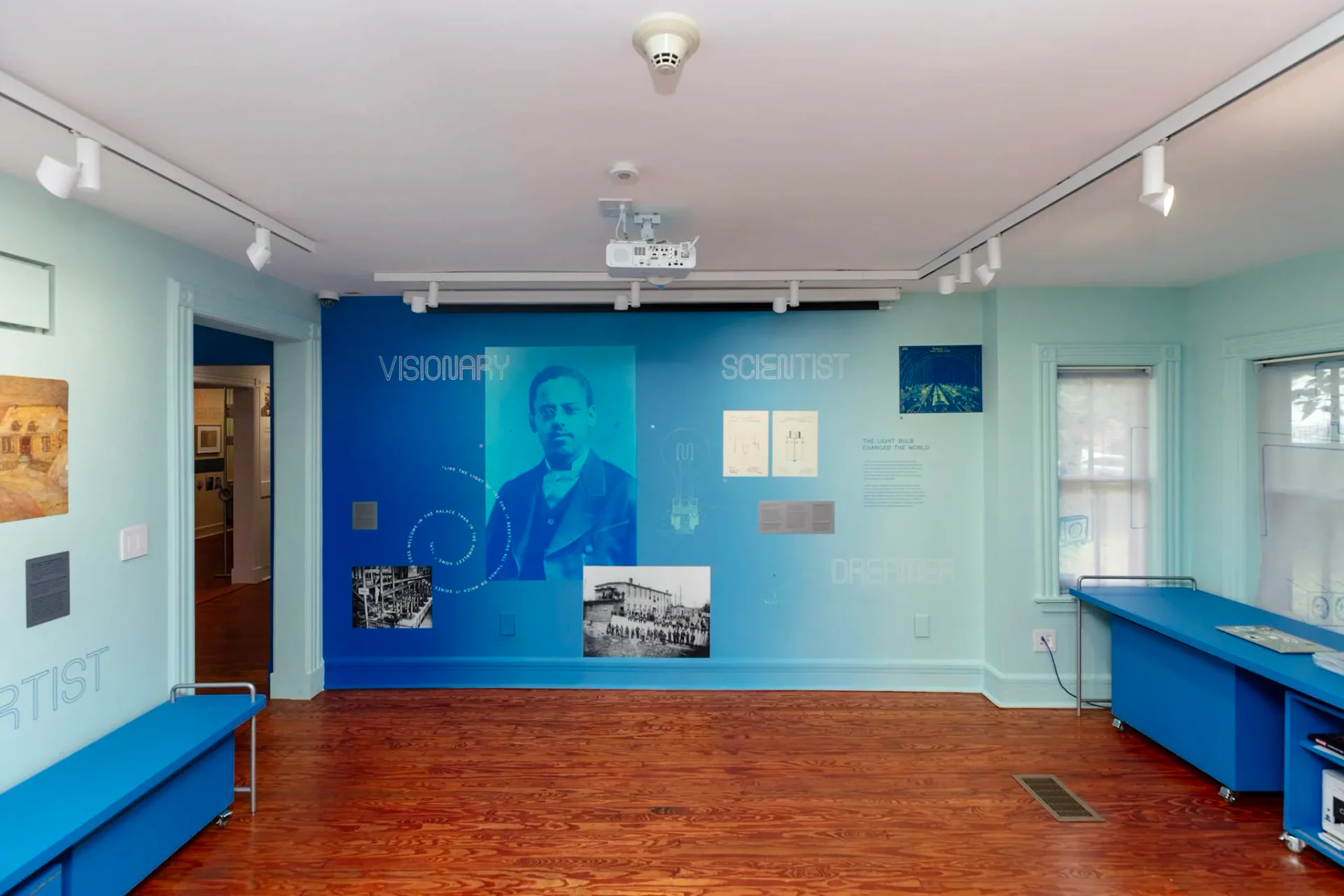 Interior gallery space with mint-green walls, blue bench seating, and a large blue wall display featuring portraits and text panels under track lighting.