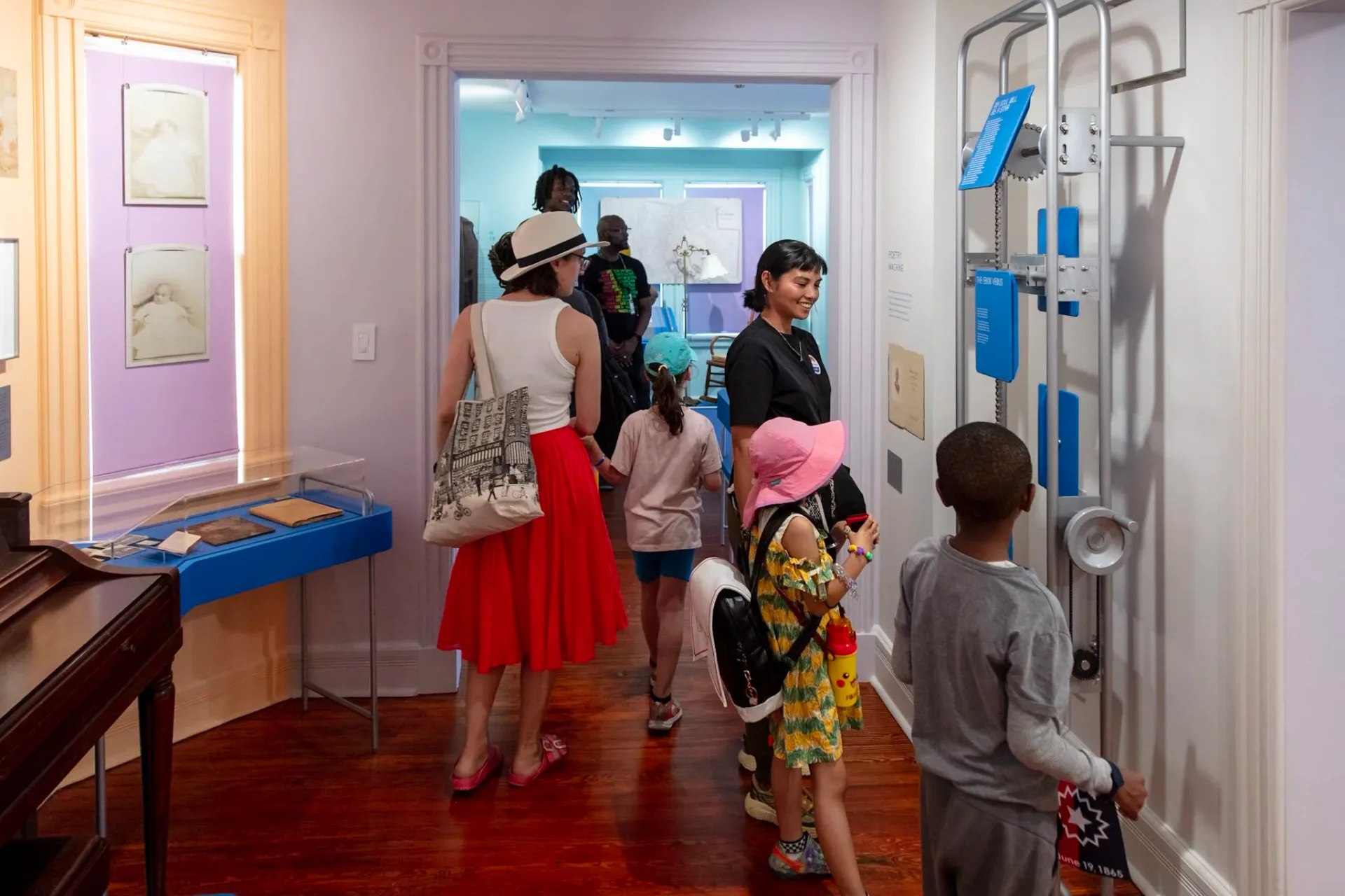 Museum gallery interior with visitors exploring exhibits, featuring purple and blue wall displays, wooden flooring, and interactive display cases.