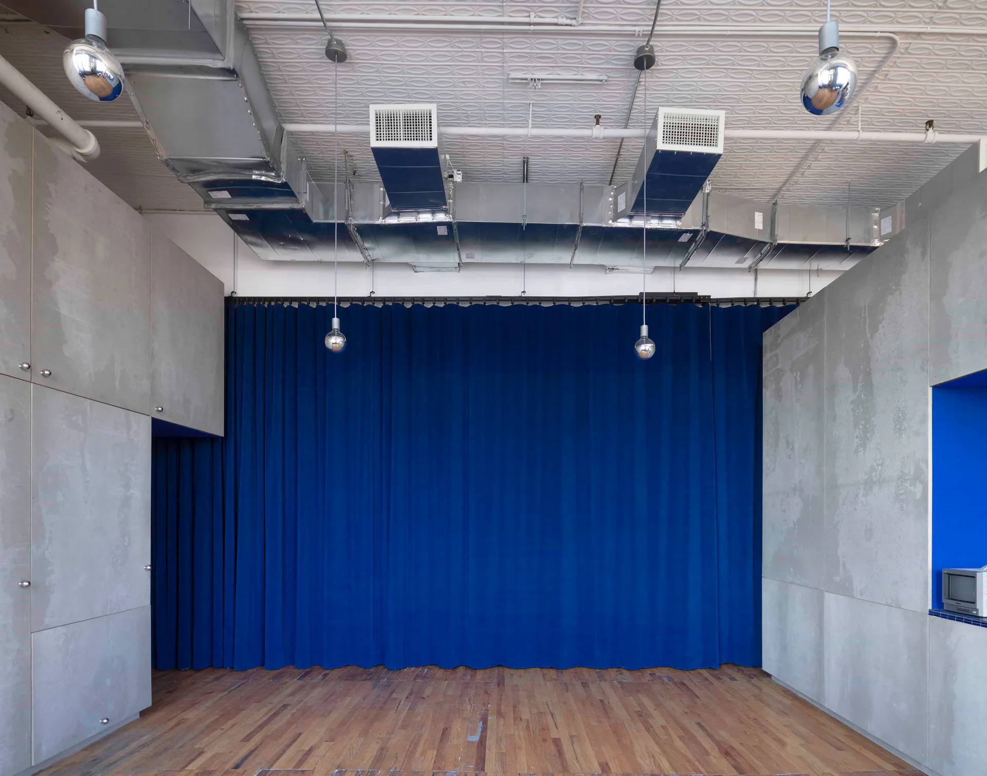 Interior of a screening room with concrete side walls, a blue curtain covering the front wall, wood flooring, and hanging globe lights from a textured white ceiling.