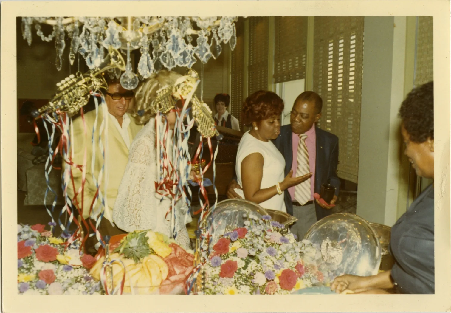 Vintage color photograph of people gathered around a decorated display with white lace, gold ornaments, and floral arrangements indoors.