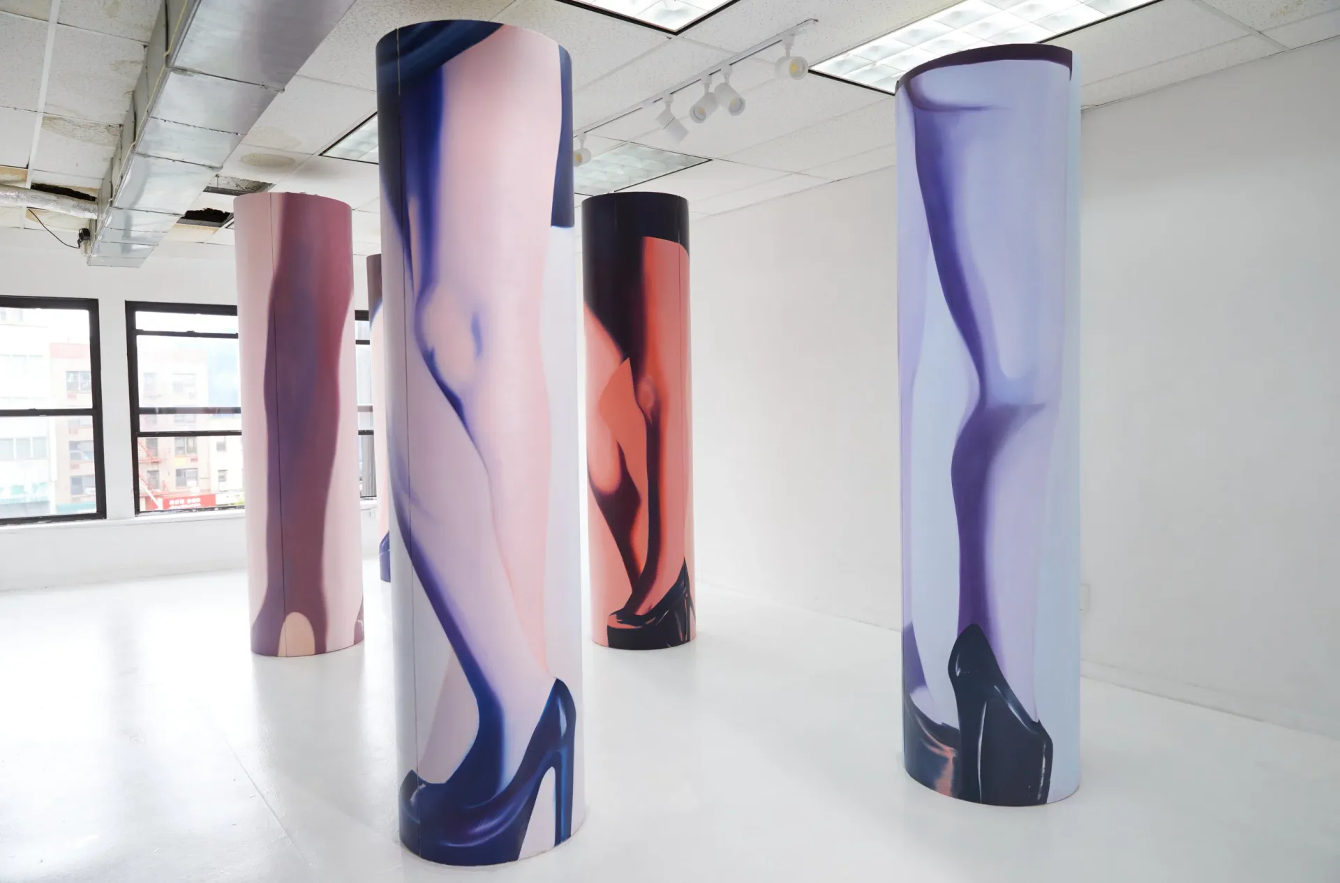 Four tall cylindrical sculptures with photographic prints of legs in high heels, featuring gradient color transitions from pink to purple to navy, installed in a white gallery space.
