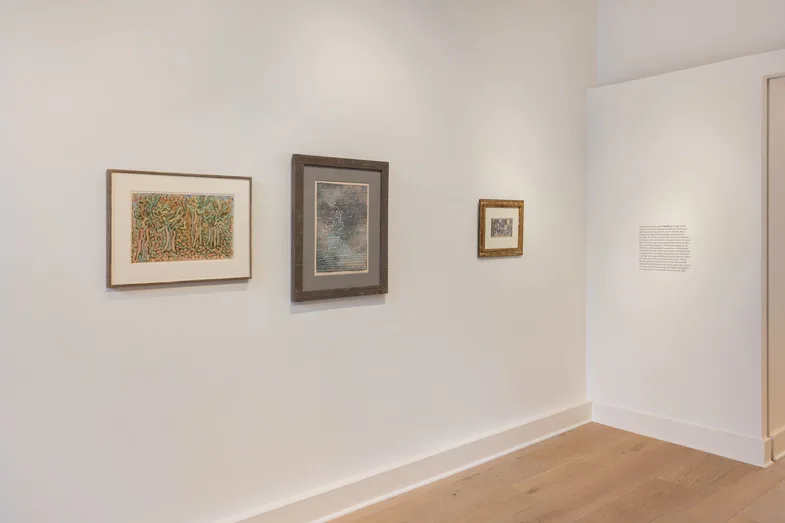 Gallery corner space with light wood floor, white walls displaying four framed works of varying sizes, including colorful and monochromatic pieces.